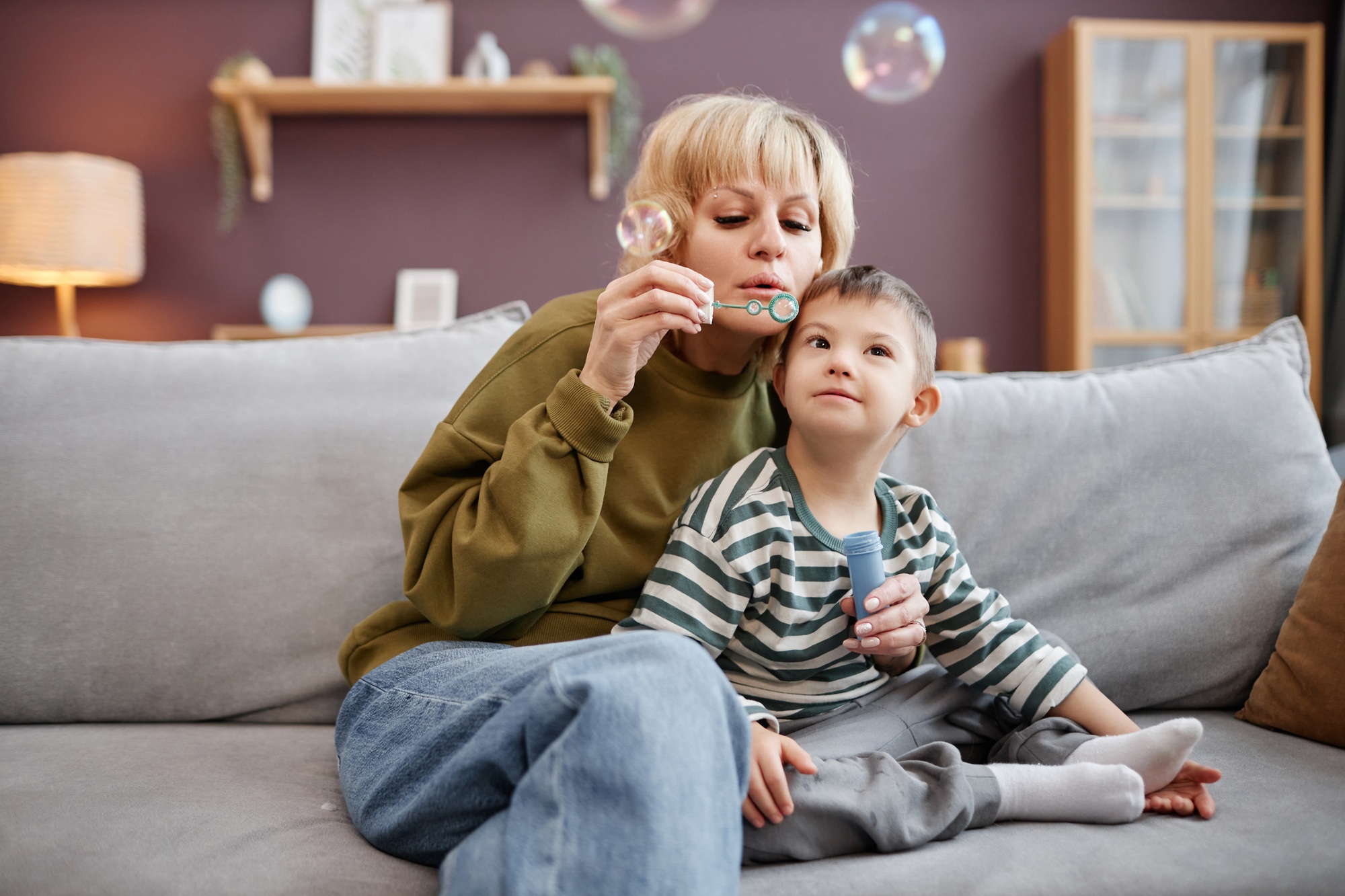Mother and child with down syndrome blowing bubbles sitting on sofa at home
